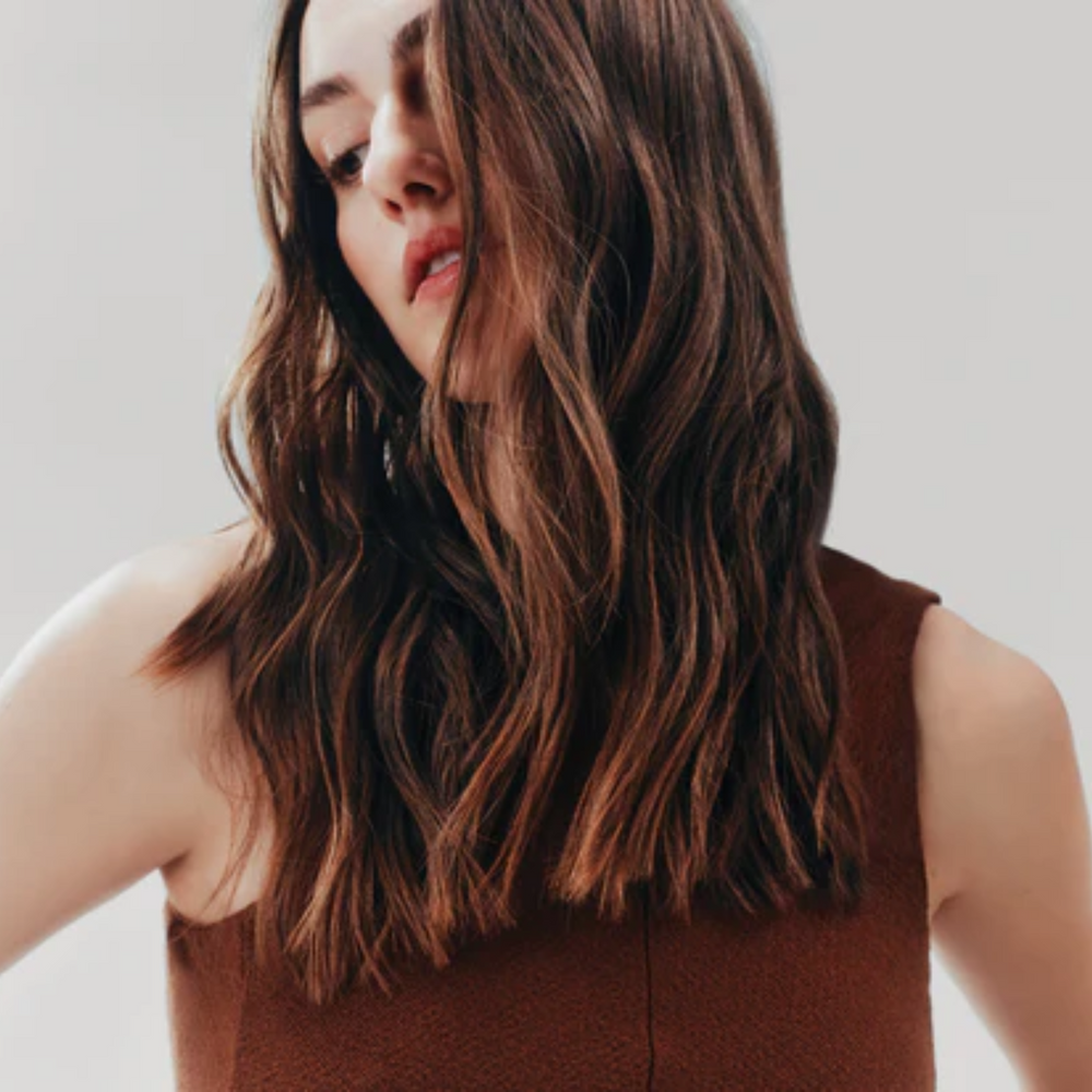 Spray AG Care Tousled Texture Body & Shine posé sur fond neutre, cheveux légèrement ondulés en arrière-plan