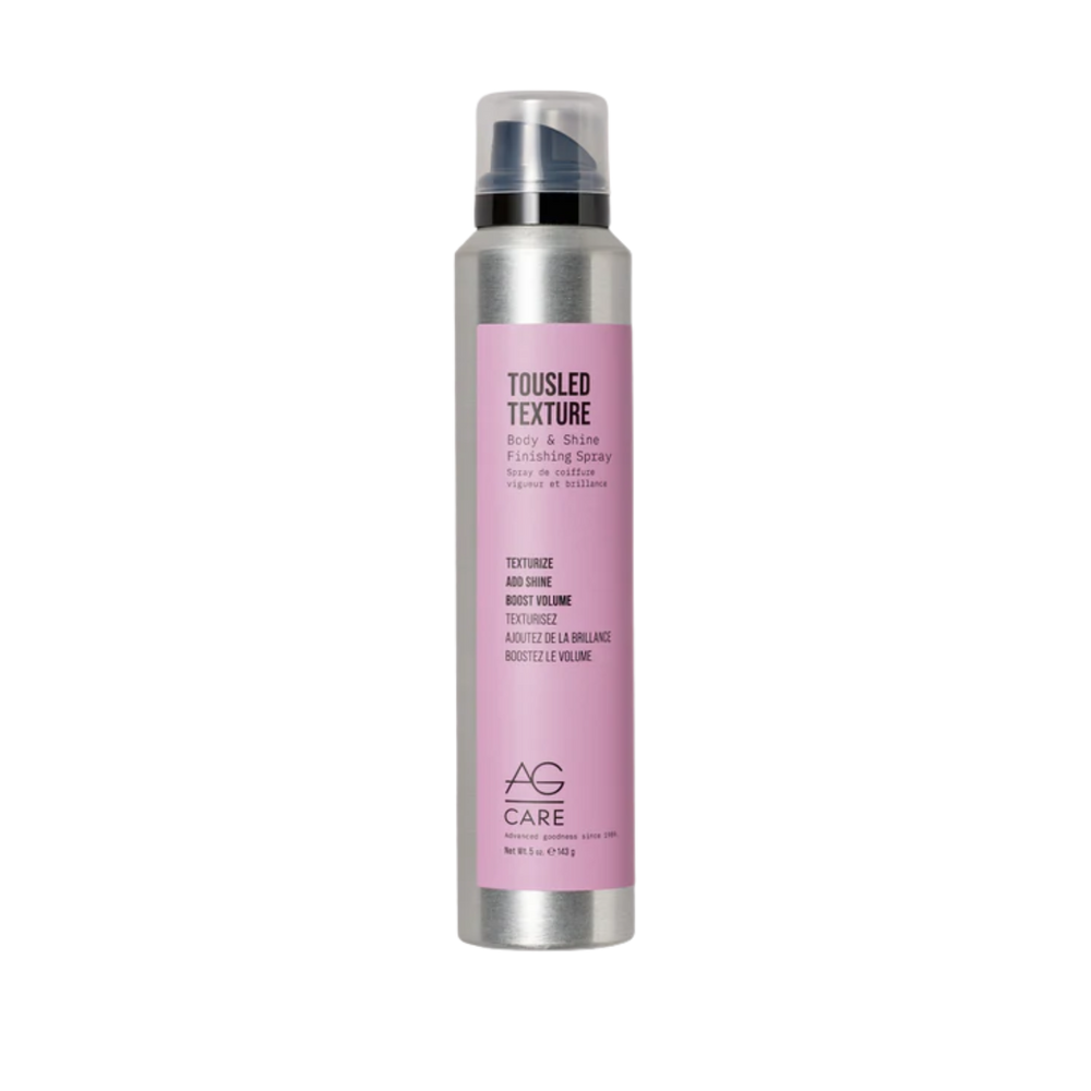 Spray AG Care Tousled Texture Body & Shine posé sur fond neutre, cheveux légèrement ondulés en arrière-plan